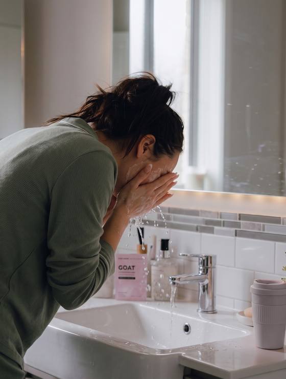 Washing her face at the bathroom sink, the GOAT pack on the vanity